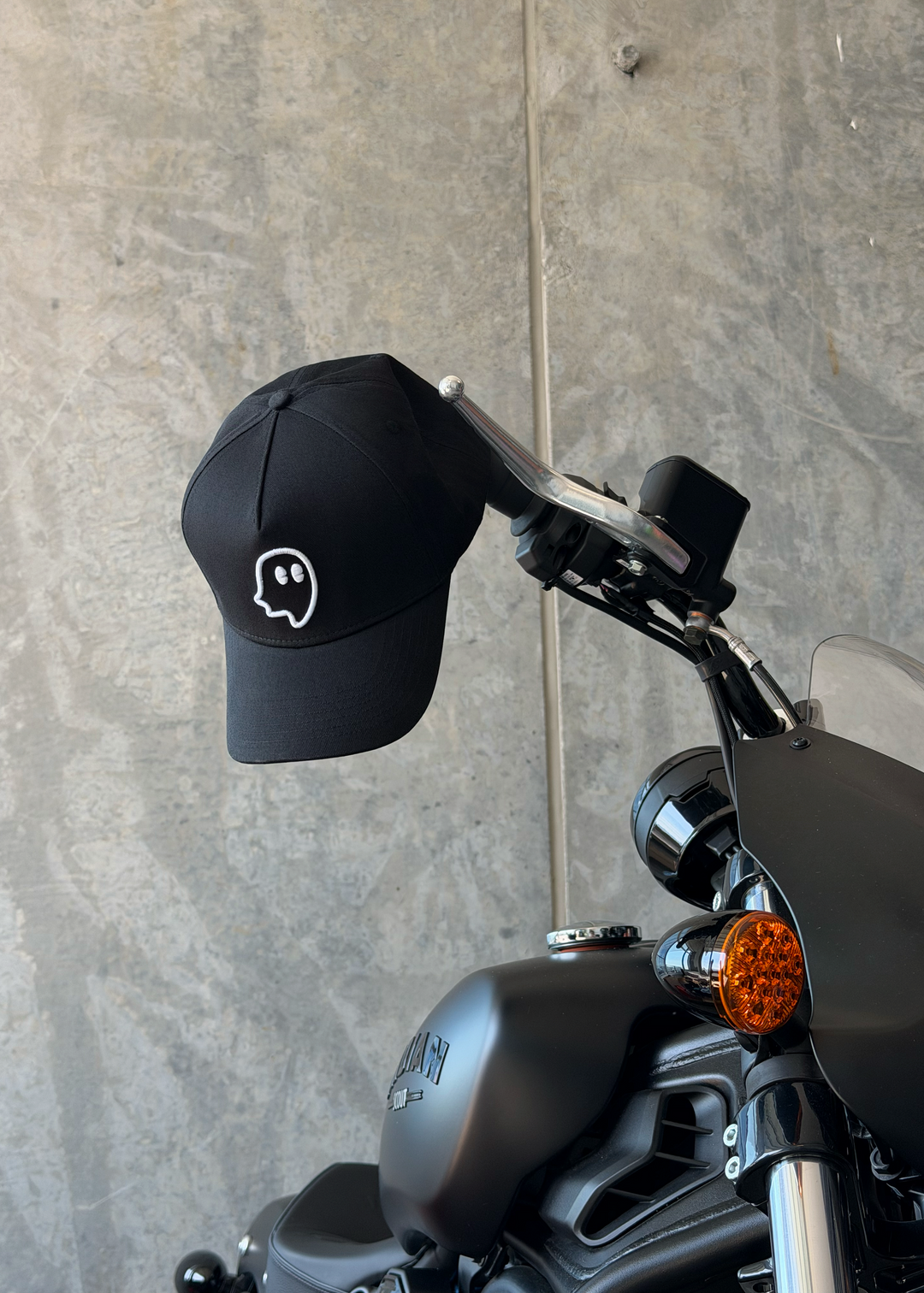 A black baseball cap with a white embroidered ghost logo hanging from a motorcycle handlebar against a textured gray concrete wall.