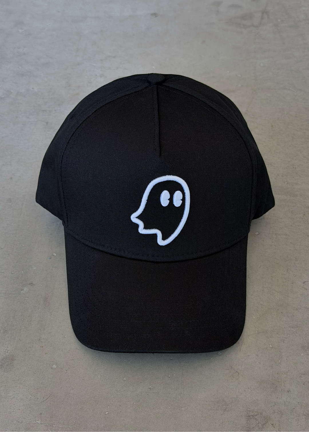 A black baseball cap with a white outline ghost graphic on the front, displayed flat on a textured gray concrete surface.