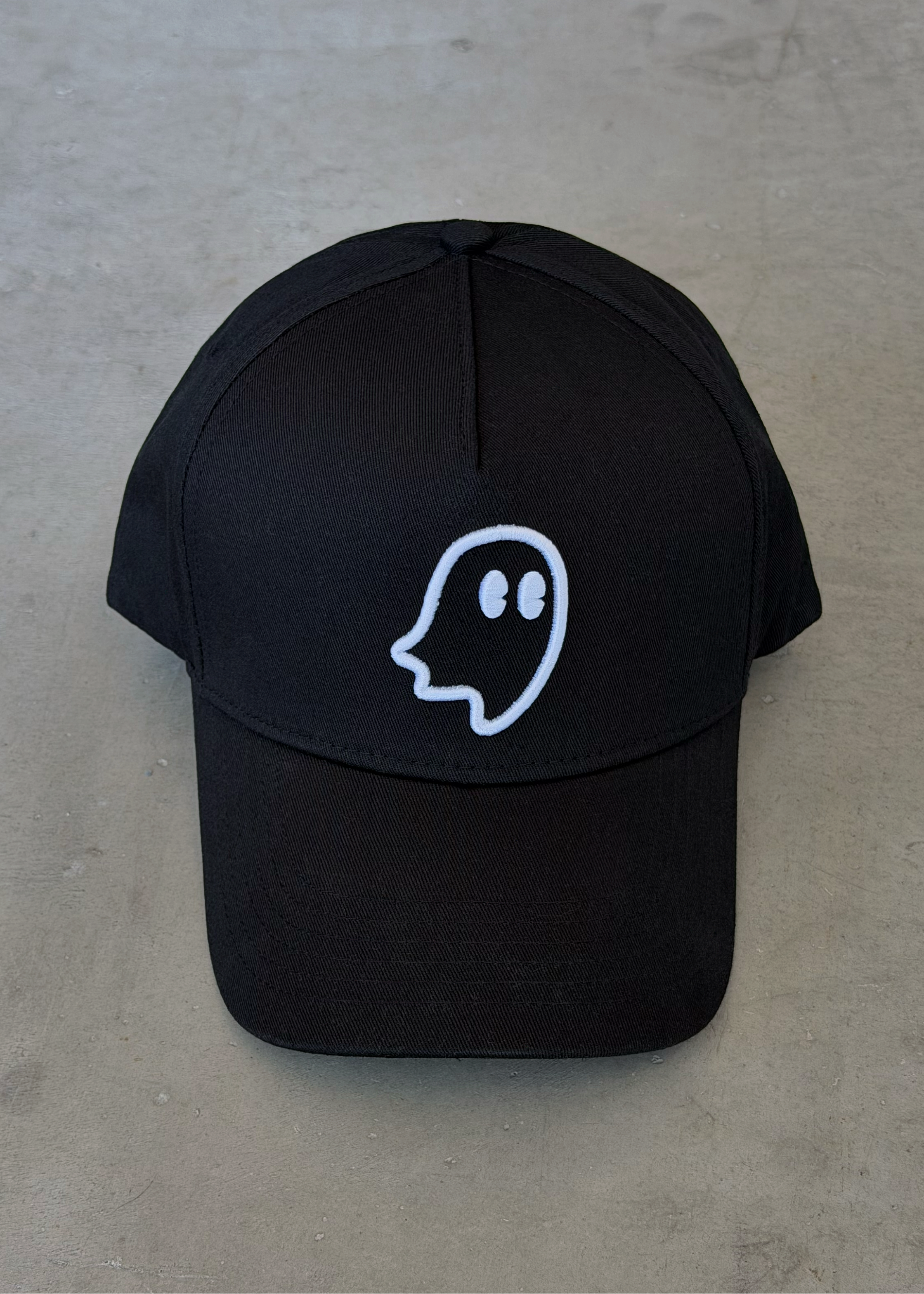 A black baseball cap with a white outline ghost graphic on the front, displayed flat on a textured gray concrete surface.