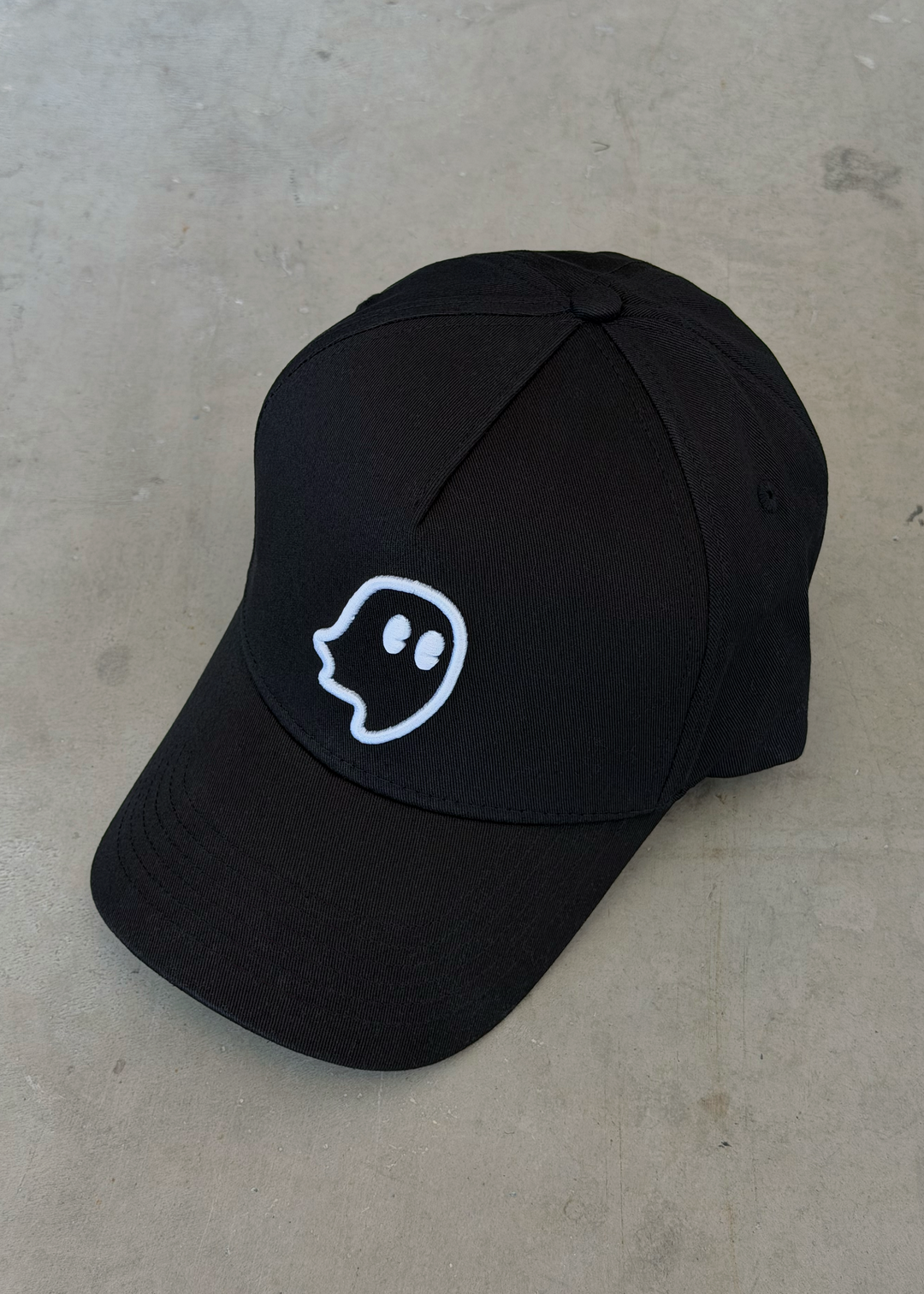 A black baseball cap with a white embroidered ghost outline logo on the front, resting on a textured gray concrete surface.