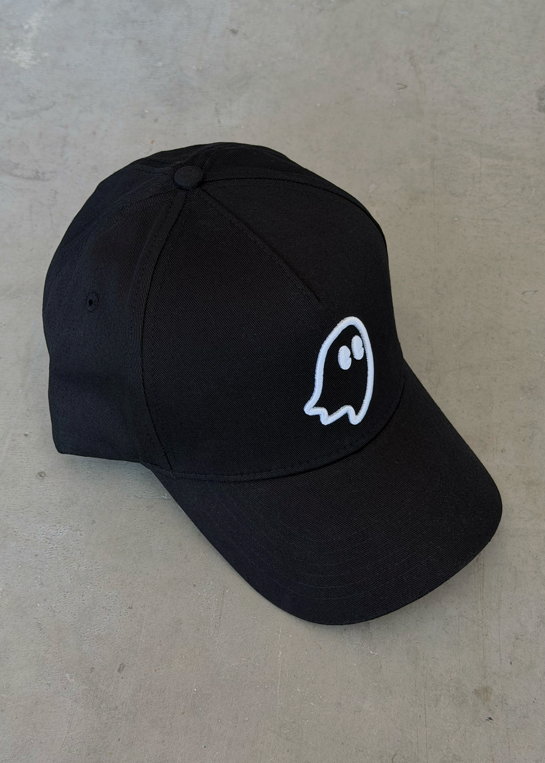 A black baseball cap with a small white embroidered ghost logo on the front, displayed on a textured gray concrete surface.