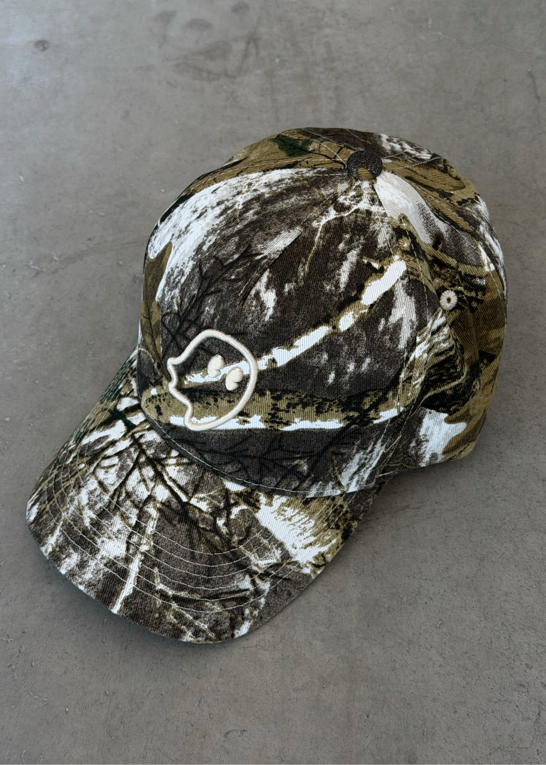 A baseball cap with a realistic brown, black, and white camouflage pattern, featuring a white embroidered deer head logo on the side, resting on a gray concrete surface.