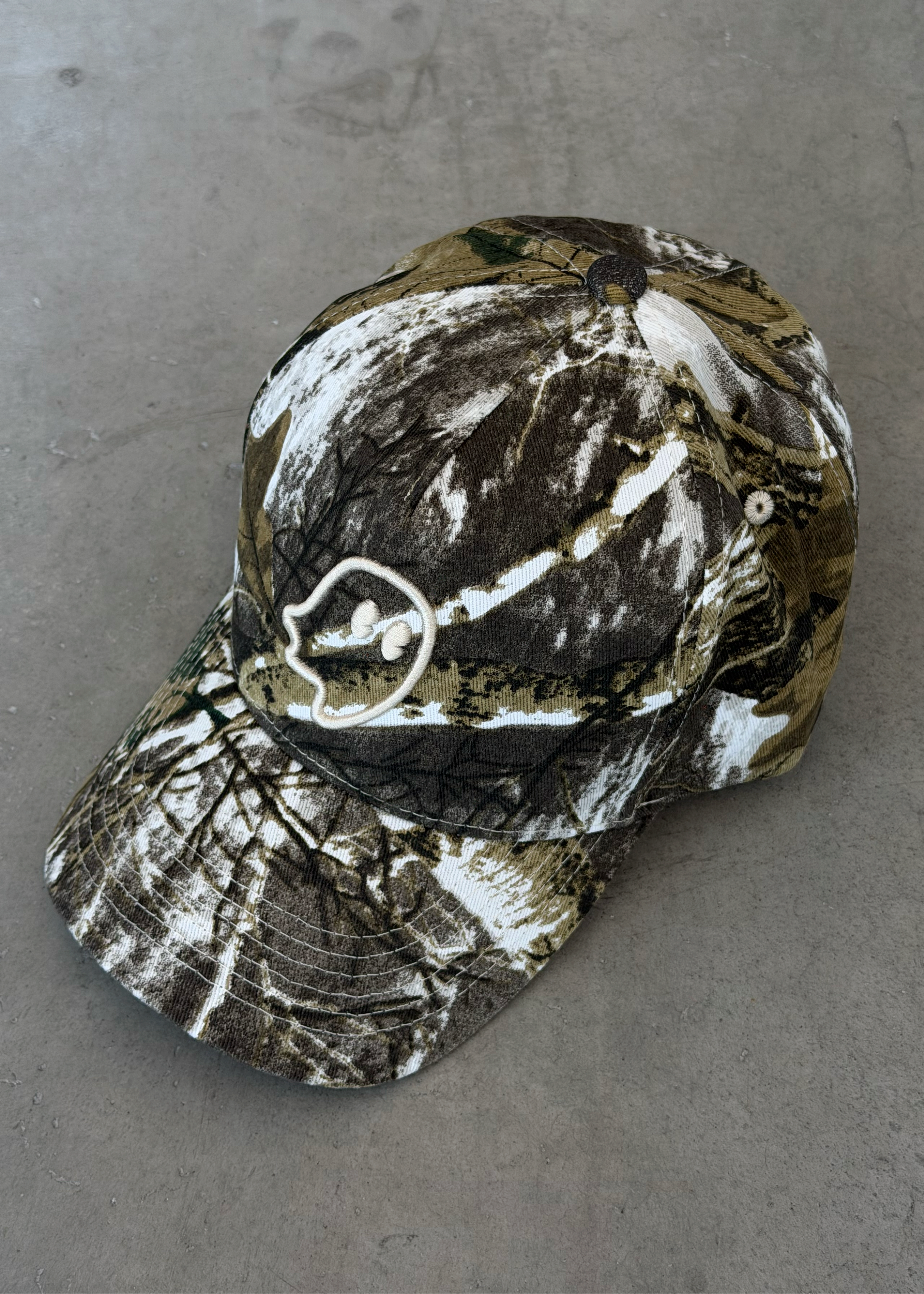 A baseball cap with a realistic brown, black, and white camouflage pattern, featuring a white embroidered deer head logo on the side, resting on a gray concrete surface.