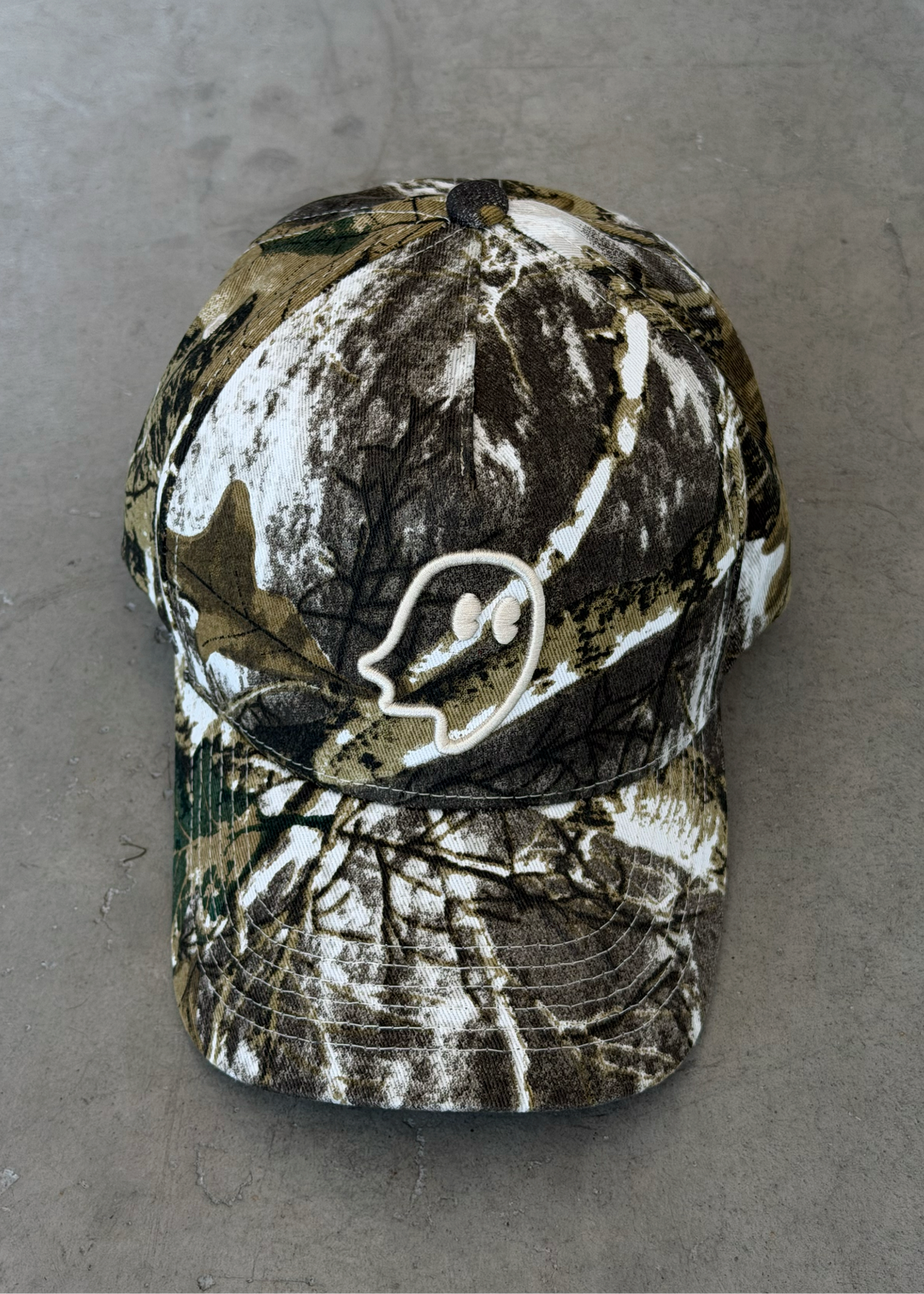 A camouflage baseball cap with a white embroidered deer head logo on the front, resting on a gray concrete floor.