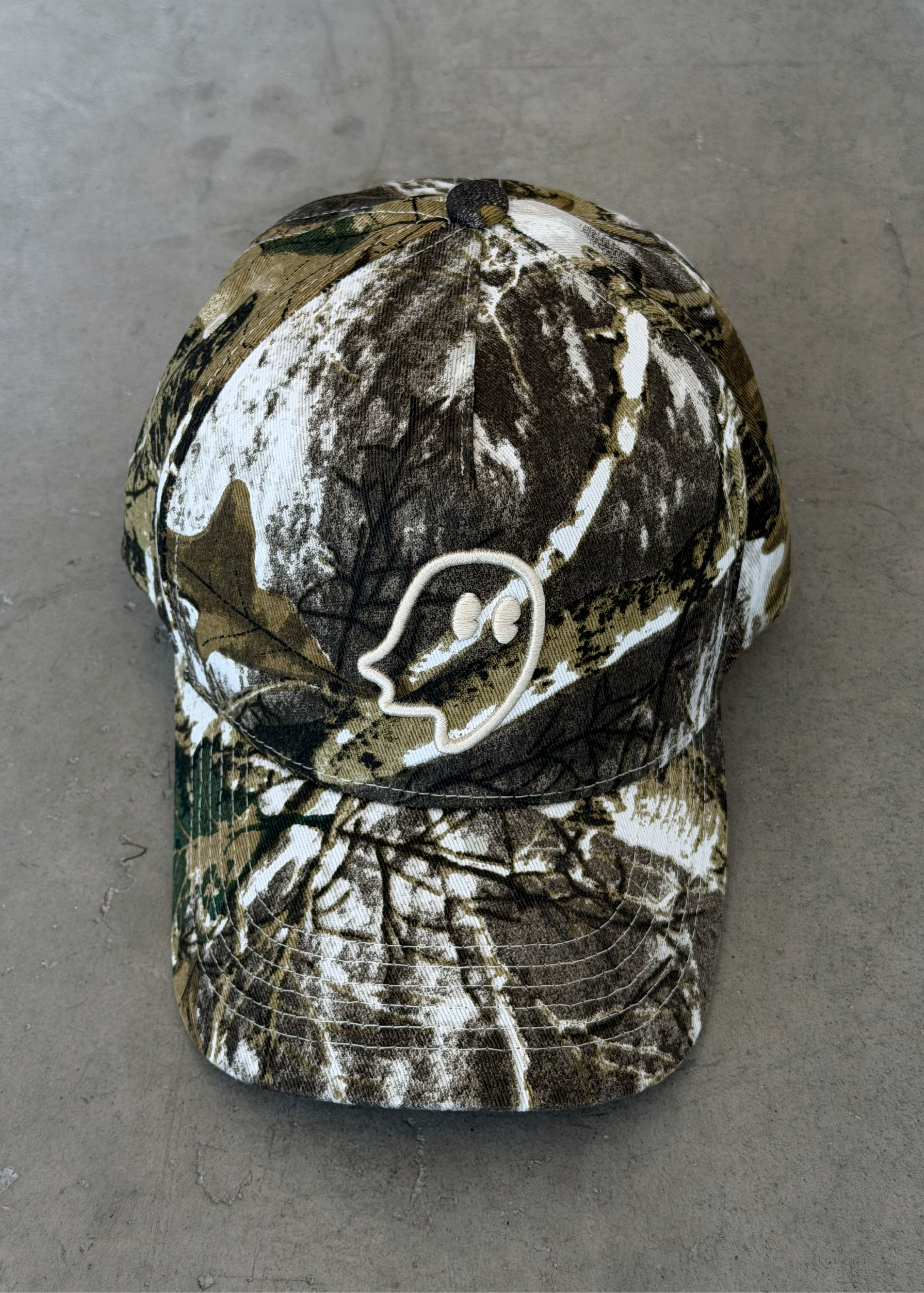 A camouflage baseball cap with a white embroidered deer head logo on the front, resting on a gray concrete floor.