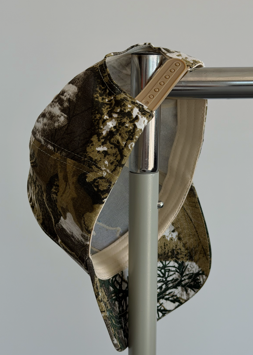 A camouflage baseball cap with a tan leather brand tab hanging from a metal stand against a plain light gray background.