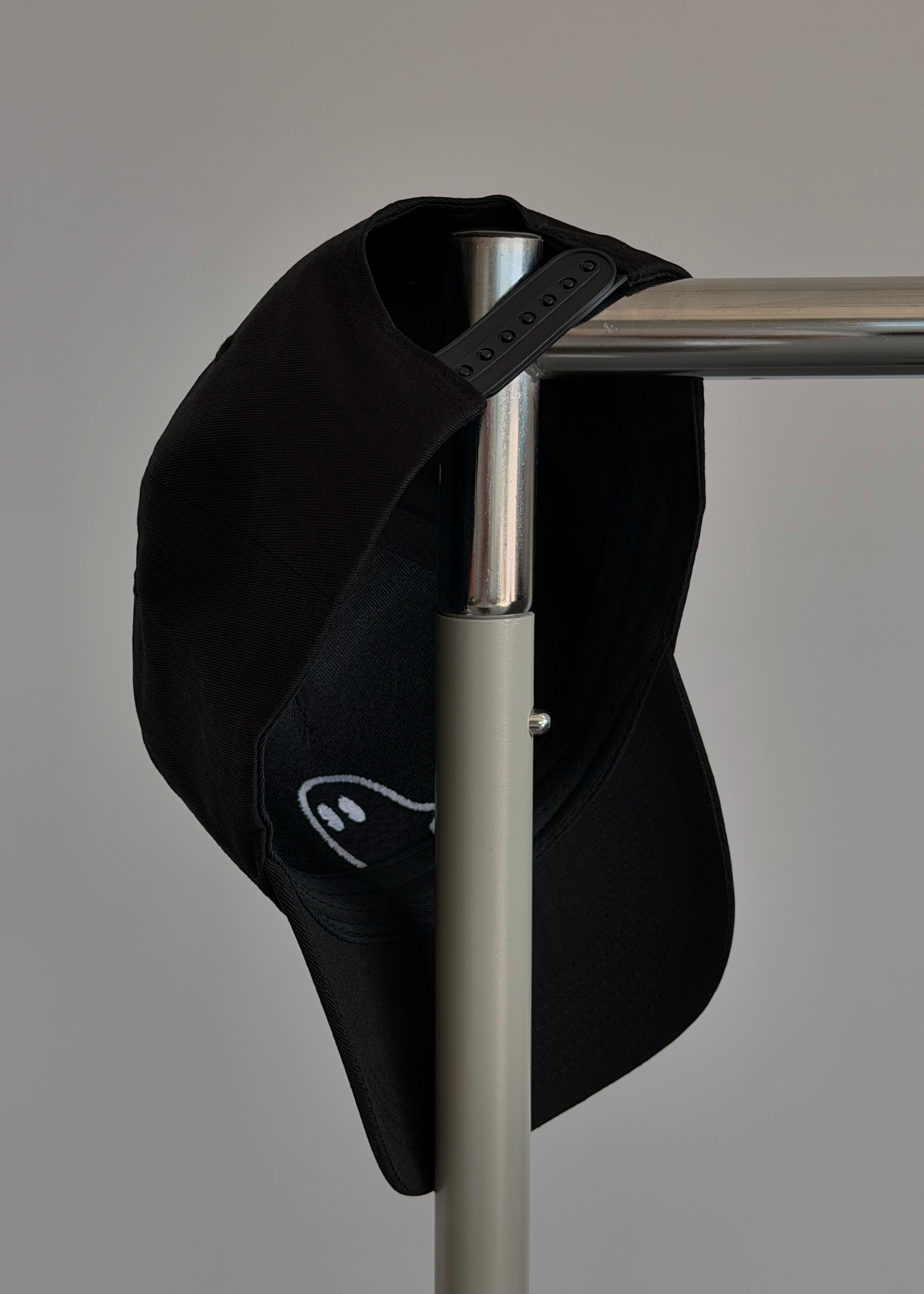 A black baseball cap with a white embroidered logo, hanging from a silver metal stand against a plain light gray background.