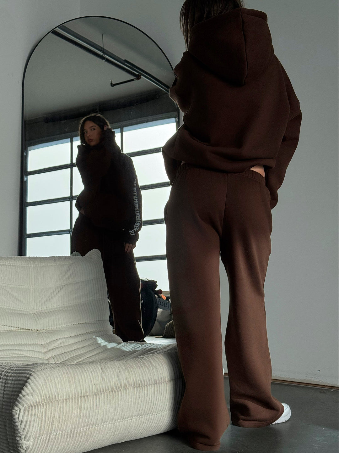 Person wearing a brown hoodie and pants standing in front of a mirror in a minimalistic room.