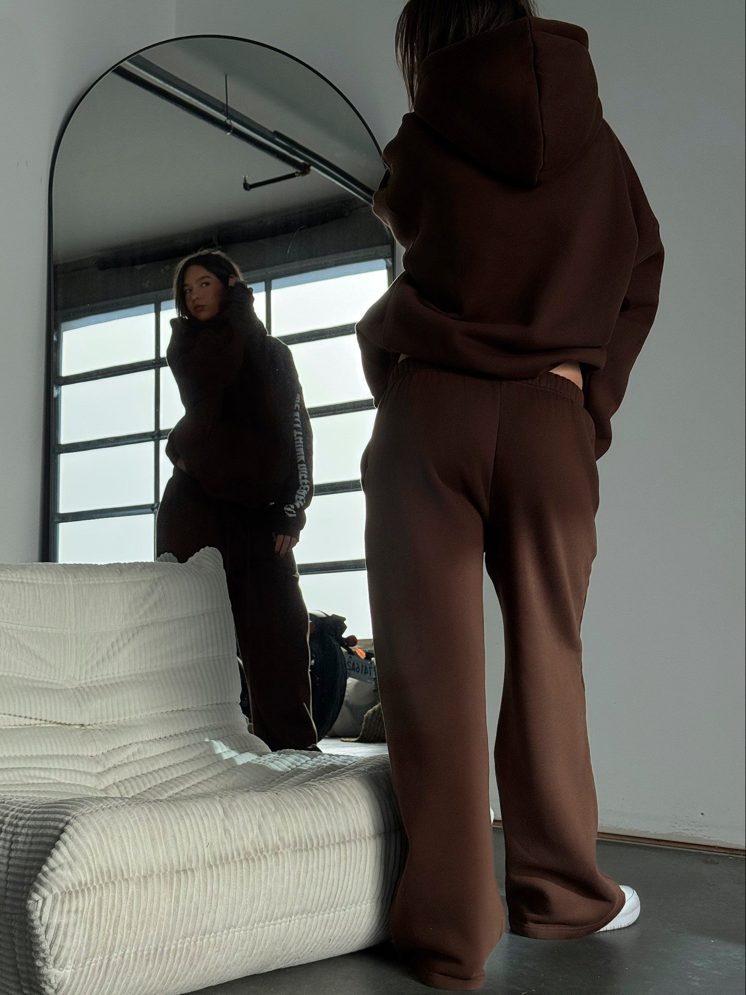 Person wearing a brown hoodie and pants standing in front of a mirror in a minimalistic room.