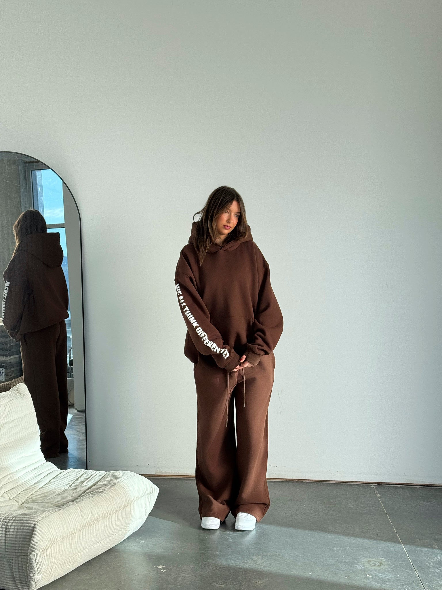 Person wearing a brown hoodie and pants standing in a minimalistic room with a mirror.