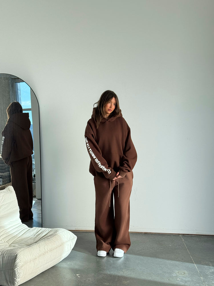 Person wearing a brown hoodie and pants standing in a minimalistic room with a mirror.