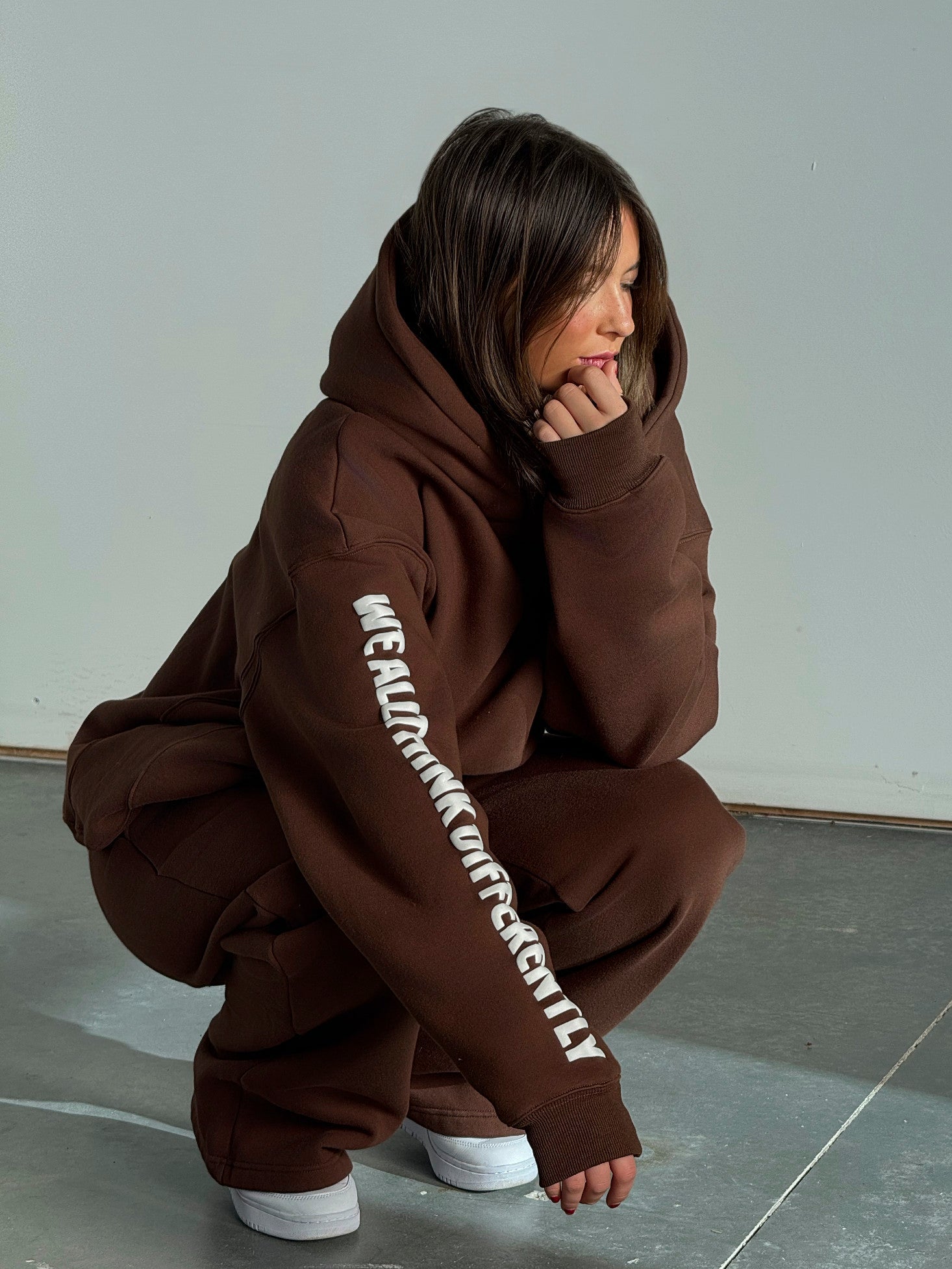 Person wearing a brown hoodie with text on the sleeve, sitting on a gray floor.