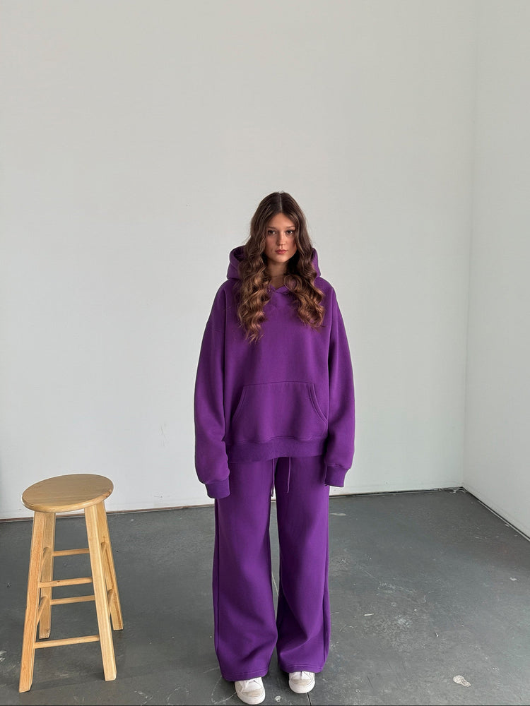 Person wearing a purple outfit standing in a minimalistic room with a wooden stool.