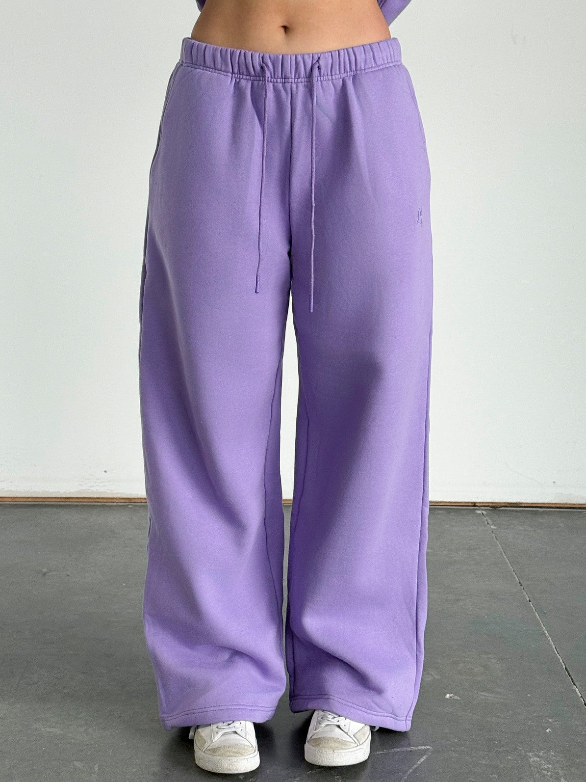 Person wearing purple sweatpants and a cropped purple hoodie against a white wall.