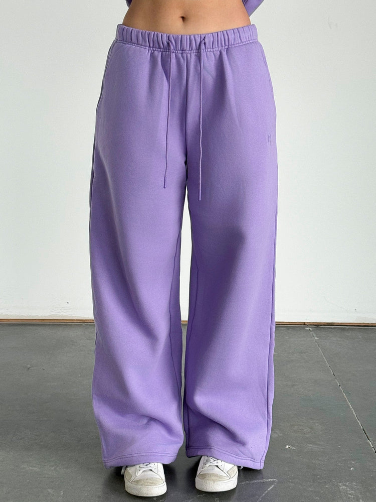 Person wearing purple sweatpants and a cropped purple hoodie against a white wall.