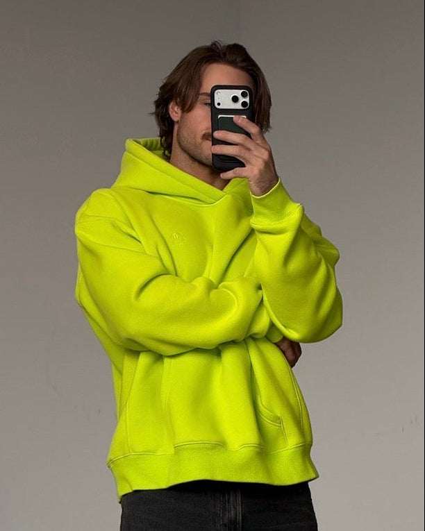 A man takes a mirror selfie wearing a bright neon yellow-green hoodie, dark gray baggy jeans, and off-white sneakers against a plain gray wall.