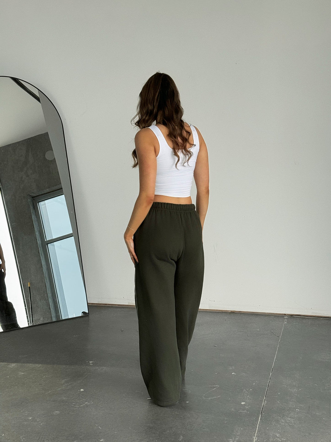 Woman wearing a white tank top and dark green pants standing in a minimalistic room with a mirror.