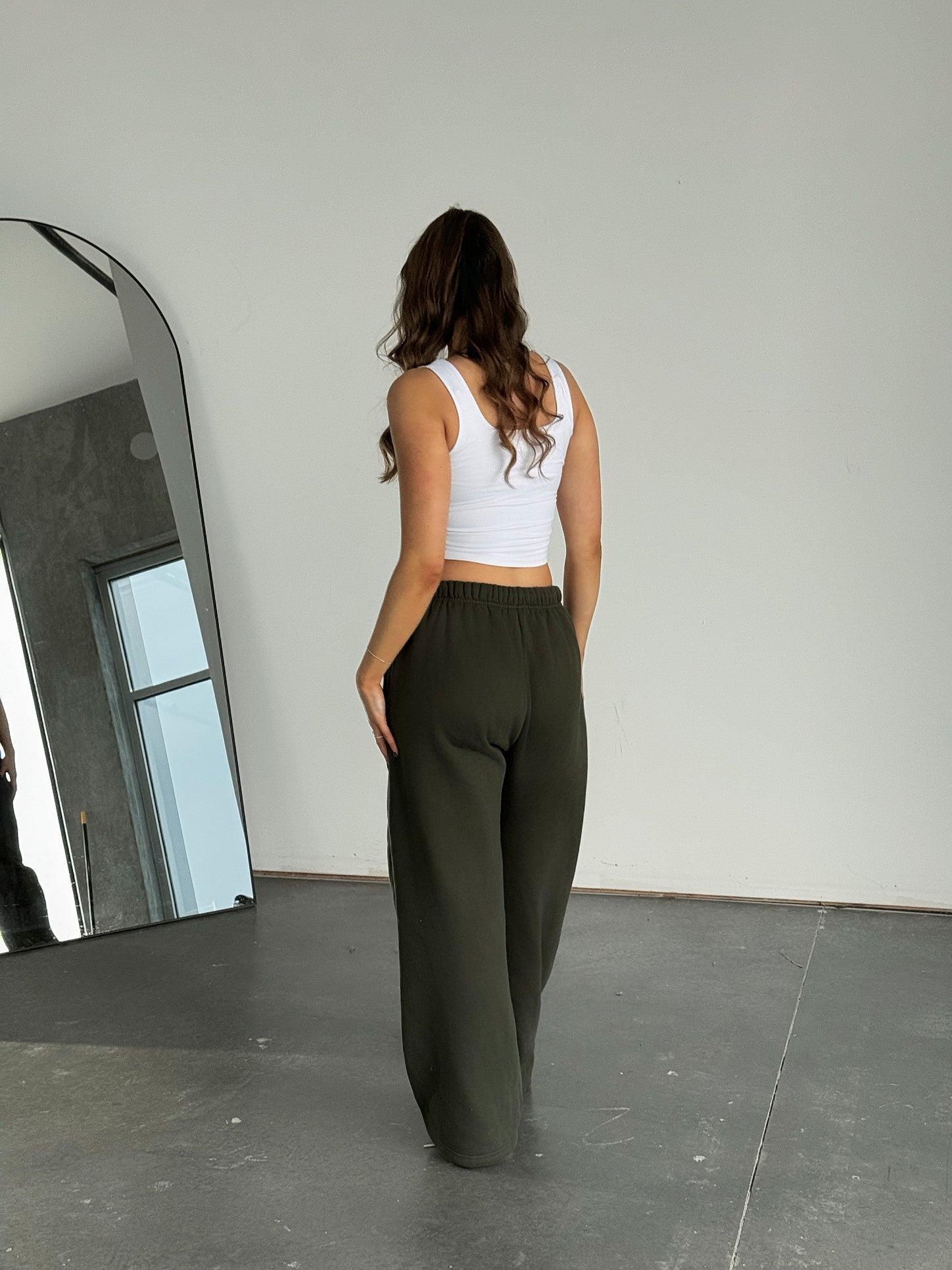 Woman wearing a white tank top and dark green pants standing in a minimalistic room with a mirror.