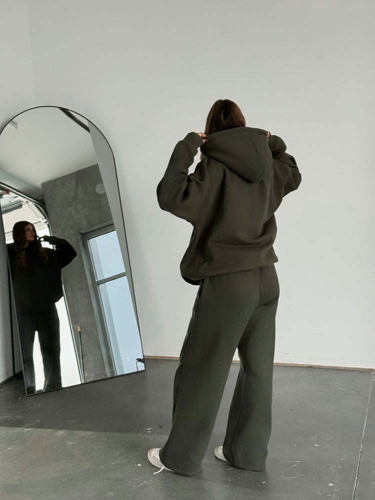 Person wearing a dark green hoodie and pants standing in front of a large mirror.