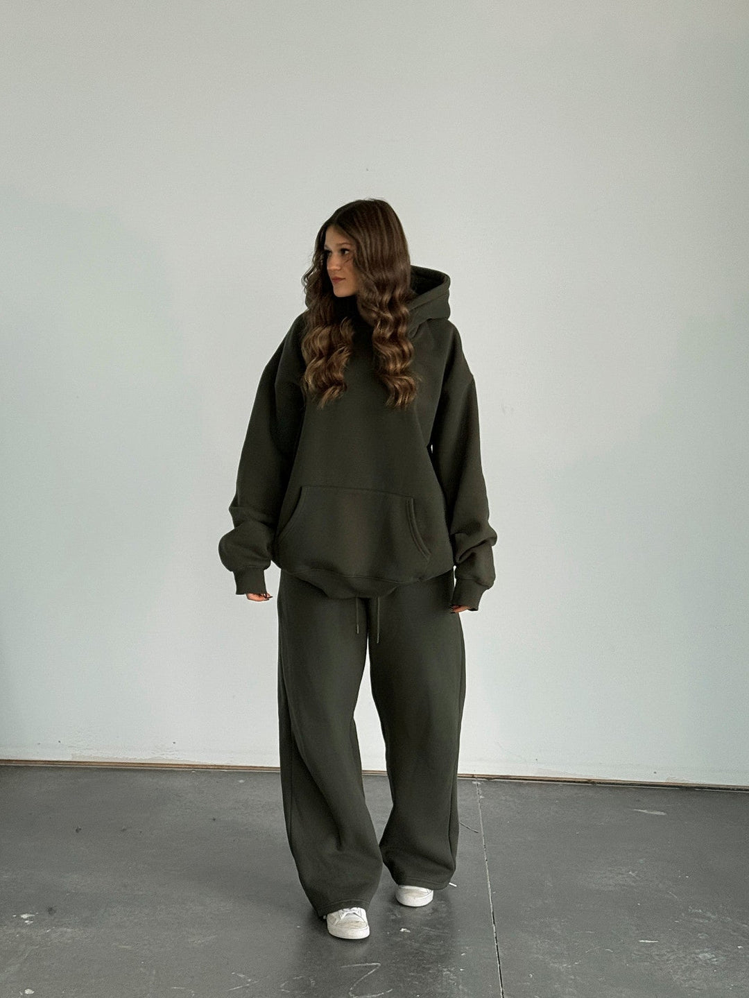 Person wearing a dark green hoodie and pants standing against a plain white wall.