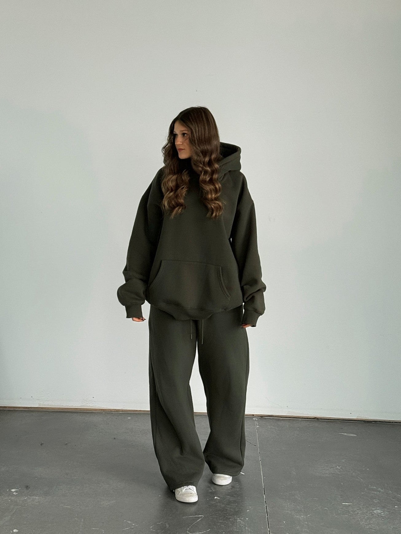 Person wearing a dark green hoodie and pants standing against a plain white wall.