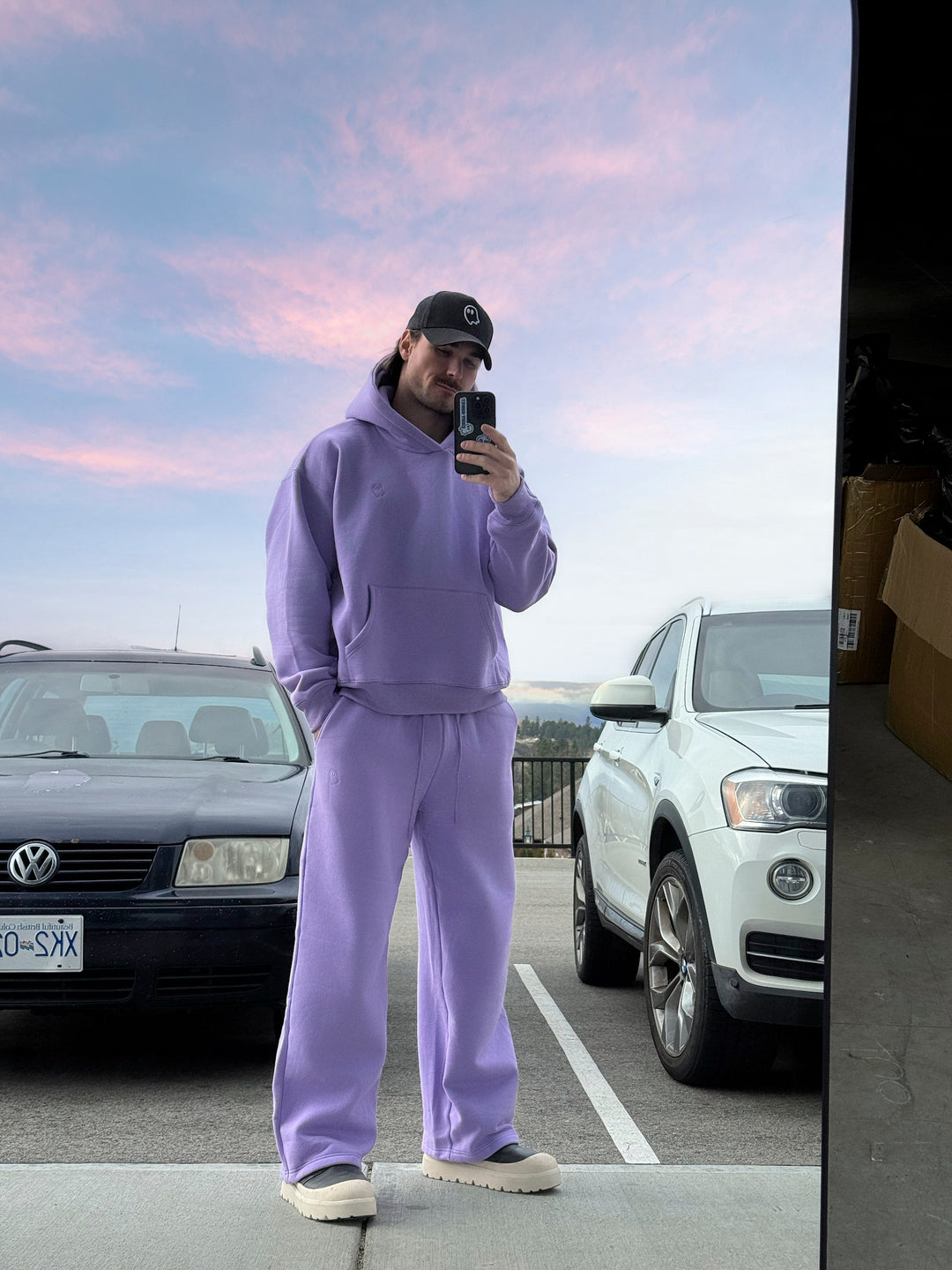 Person wearing a purple tracksuit taking a mirror selfie in a parking lot with cars and a sunset sky.