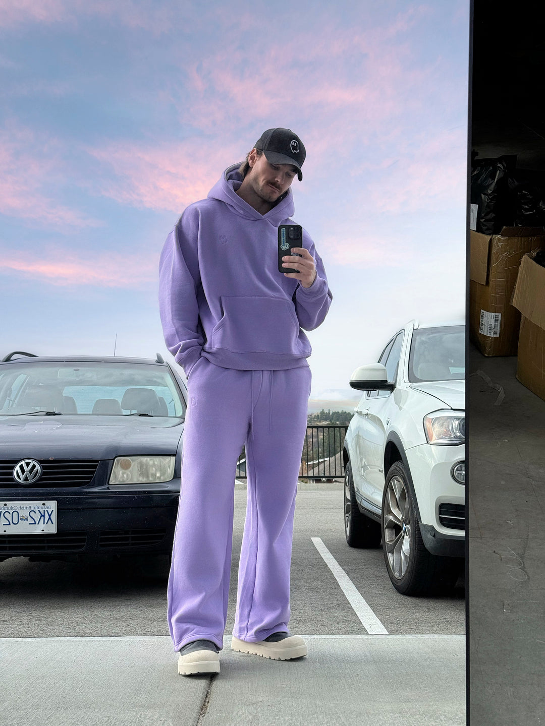 Person in a purple tracksuit taking a mirror selfie in a parking lot with cars and a pink sky.
