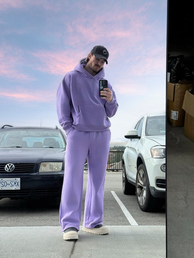 Person in a purple tracksuit taking a mirror selfie in a parking lot with cars and a pink sky.