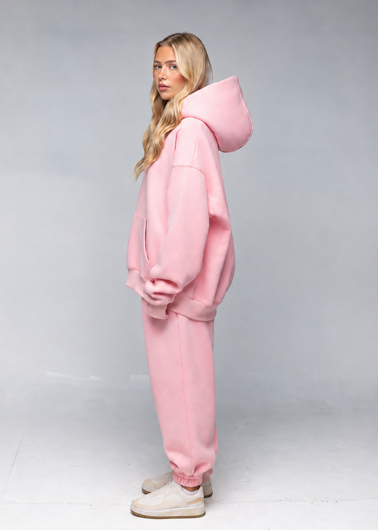 A woman modeling a light pink oversized hoodie and matching jogger sweatpants set, standing in profile against a neutral gray studio background.