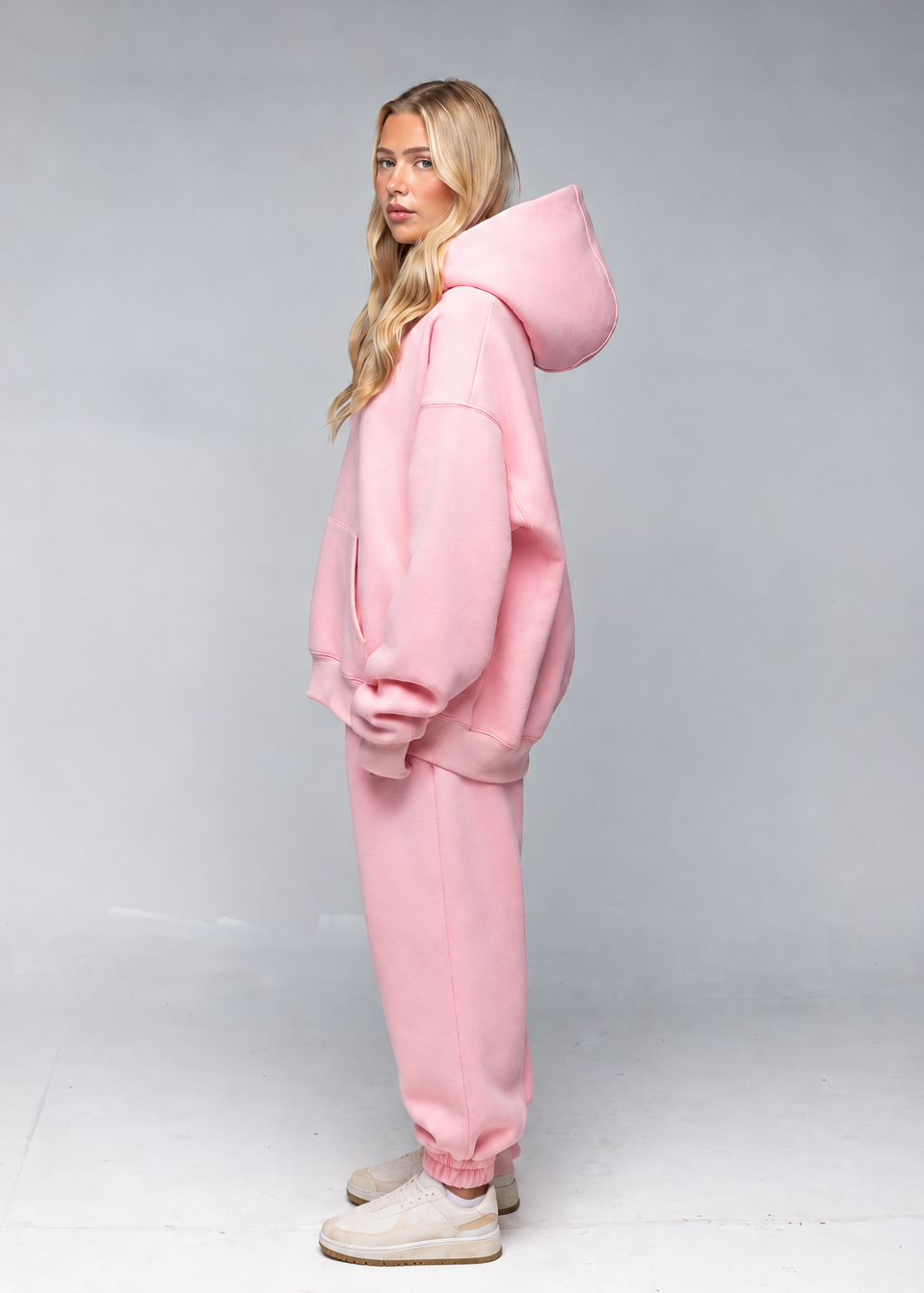 A woman modeling a light pink oversized hoodie and matching jogger sweatpants set, standing in profile against a neutral gray studio background.