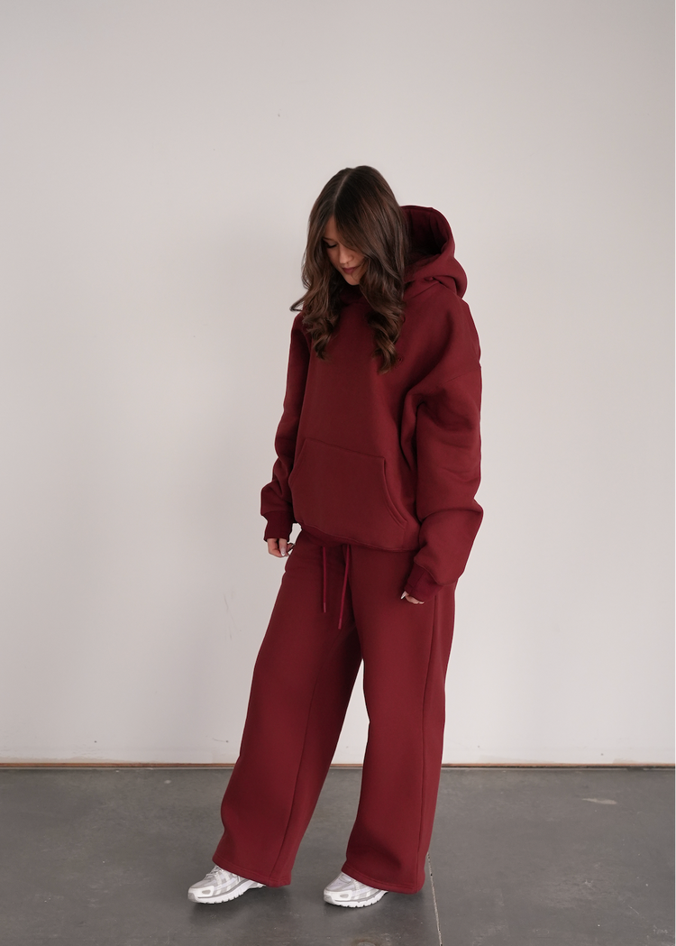 Person wearing a maroon hoodie and pants standing against a plain white wall.