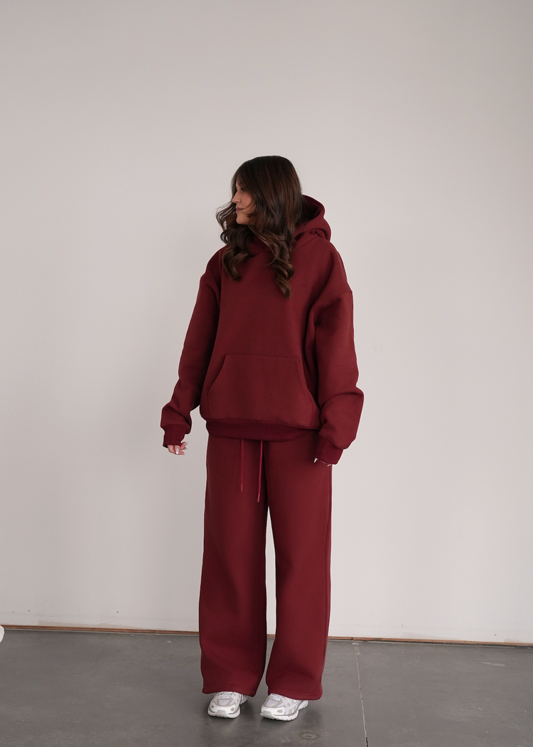 Person wearing a burgundy hoodie and pants standing against a plain white wall.