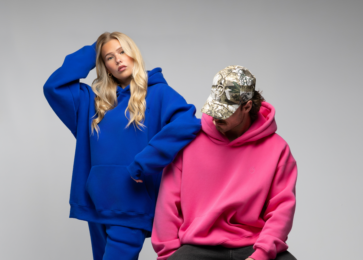 Two models posing in oversized hoodies, one wearing a vibrant royal blue hoodie and matching pants, and the other wearing a bright pink hoodie, dark pants, and a camouflage baseball cap, set against a plain gray studio background.