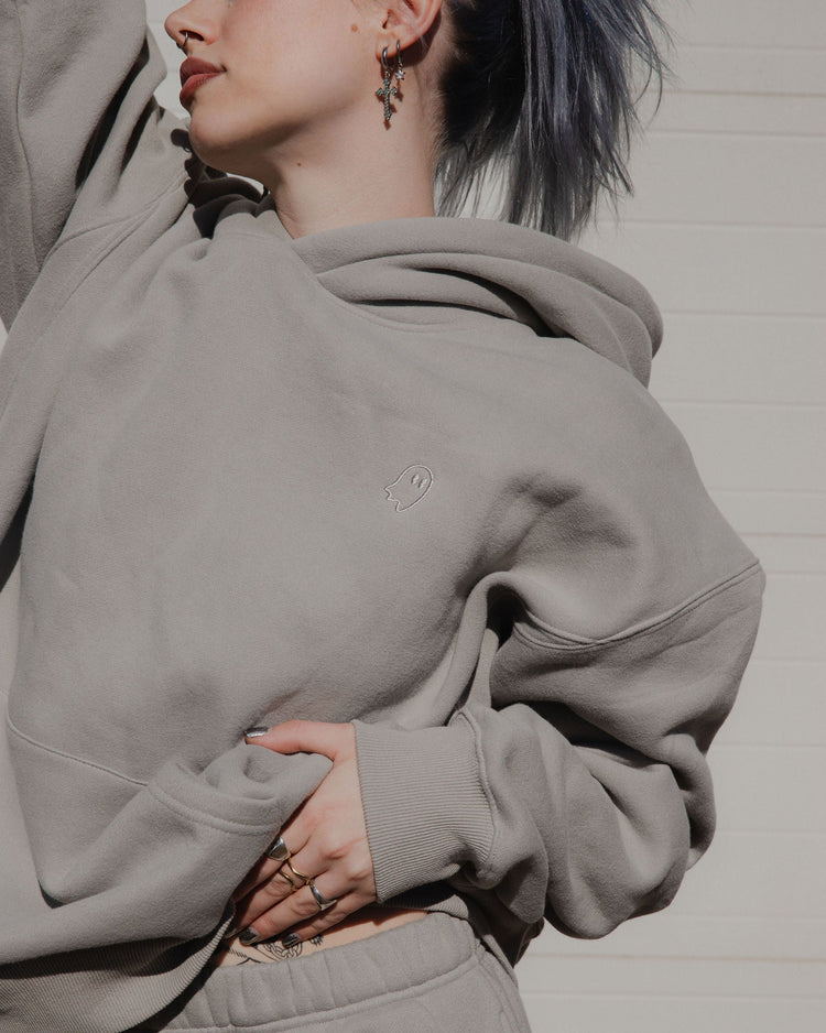 Person wearing a gray hoodie with a heart emblem, standing against a neutral background.