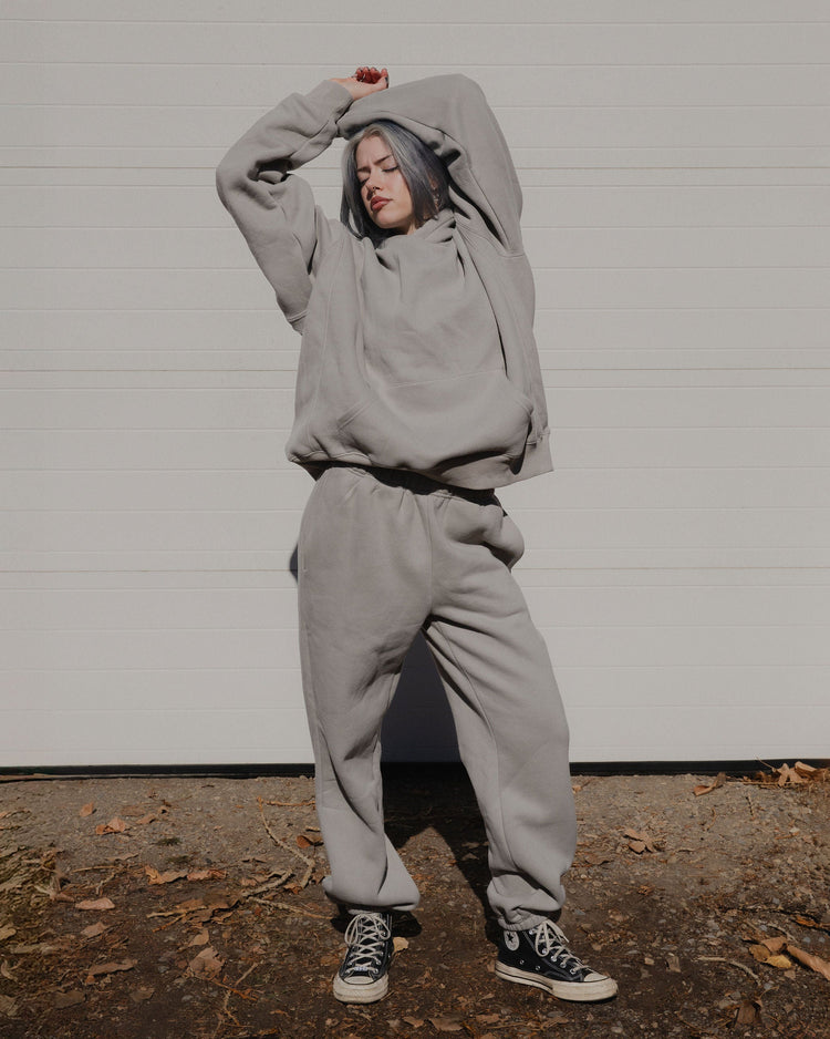 Person wearing a gray hoodie and sweatpants with black sneakers against a white wall.