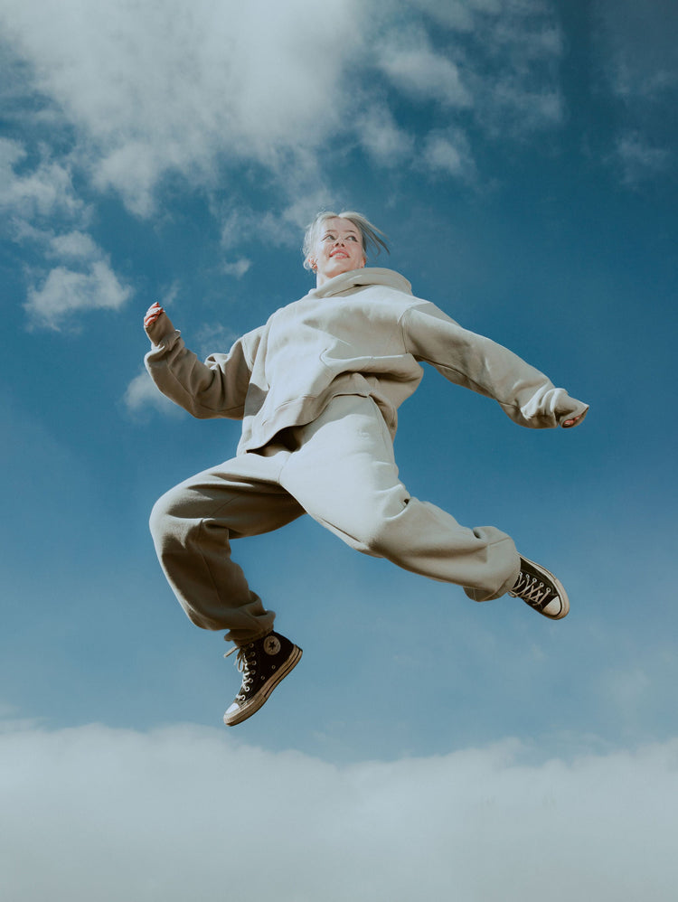 Person in beige jacket and pants jumping against a blue sky with clouds