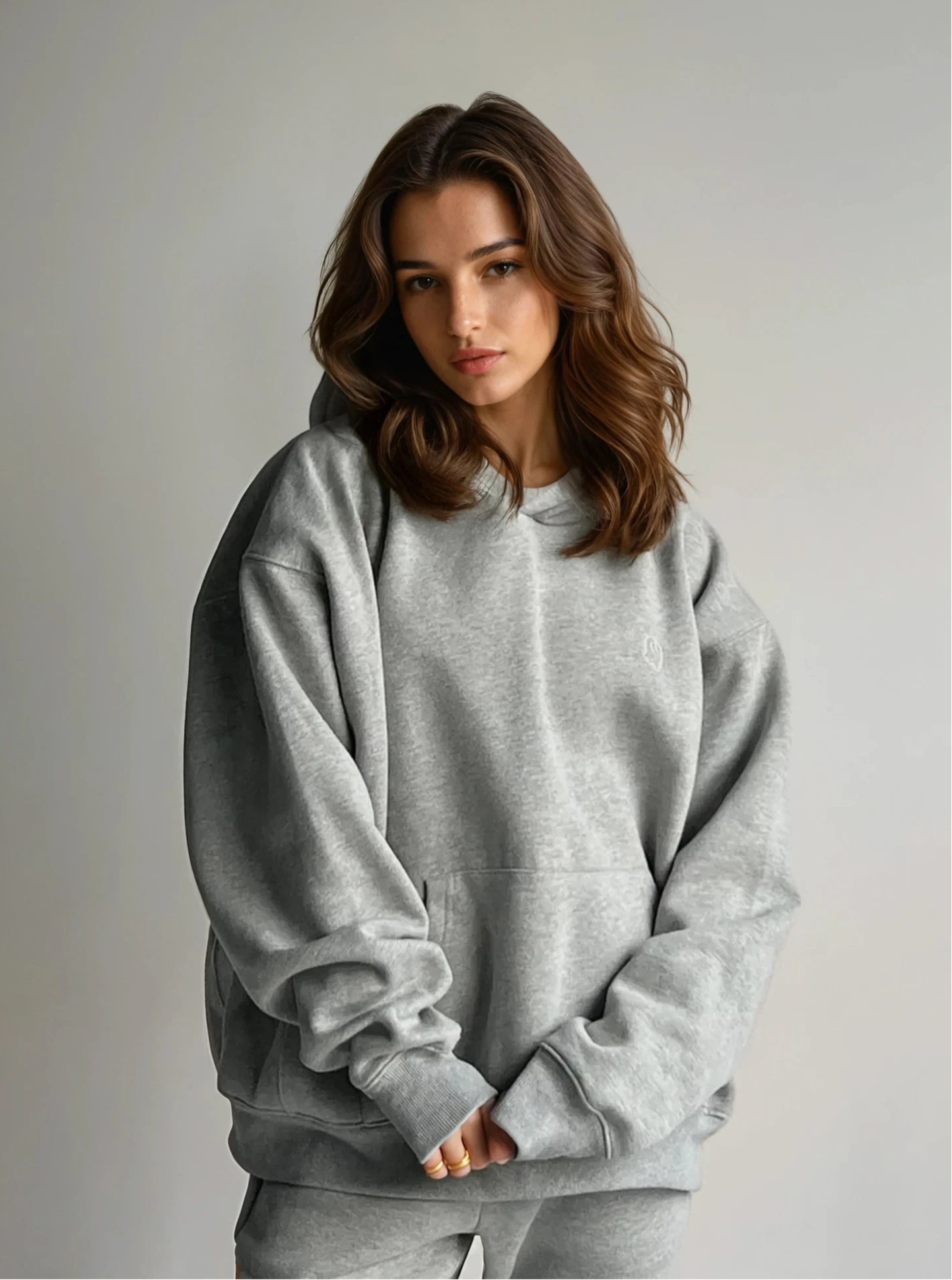 A person wearing a loose-fitting grey hoodie with a hood, standing against a grey background.